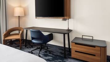 Una habitación de hotel con cama, escritorio, silla, televisor, lámpara y un mueble pequeño.