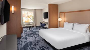 A hotel room with a large bed, a desk, a chair, a TV, and a window with a view of trees.