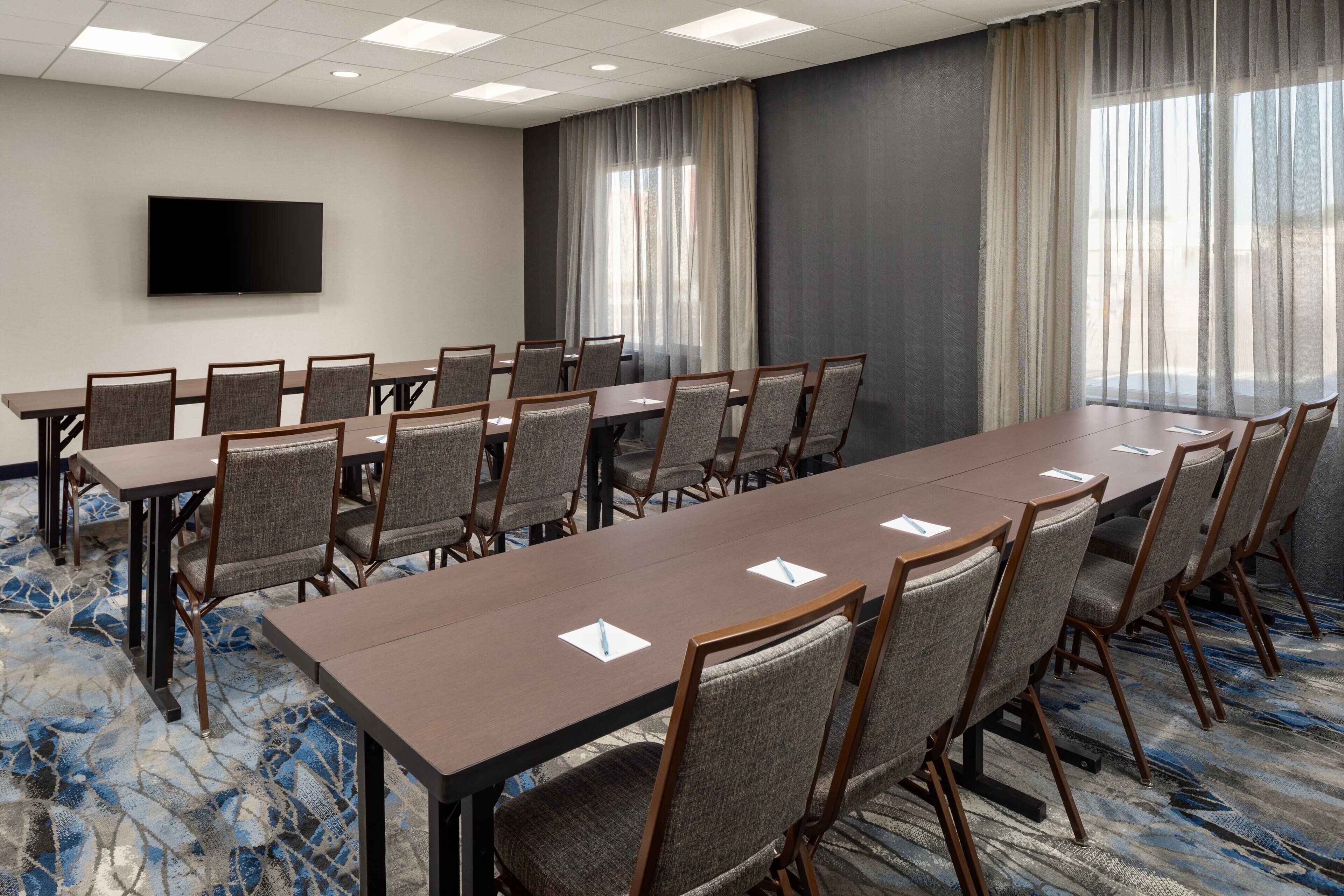 A conference room with a long table, chairs, a flat-screen TV, and large windows with curtains.