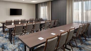 A conference room with a long table, chairs, a flat-screen TV, and large windows with curtains.