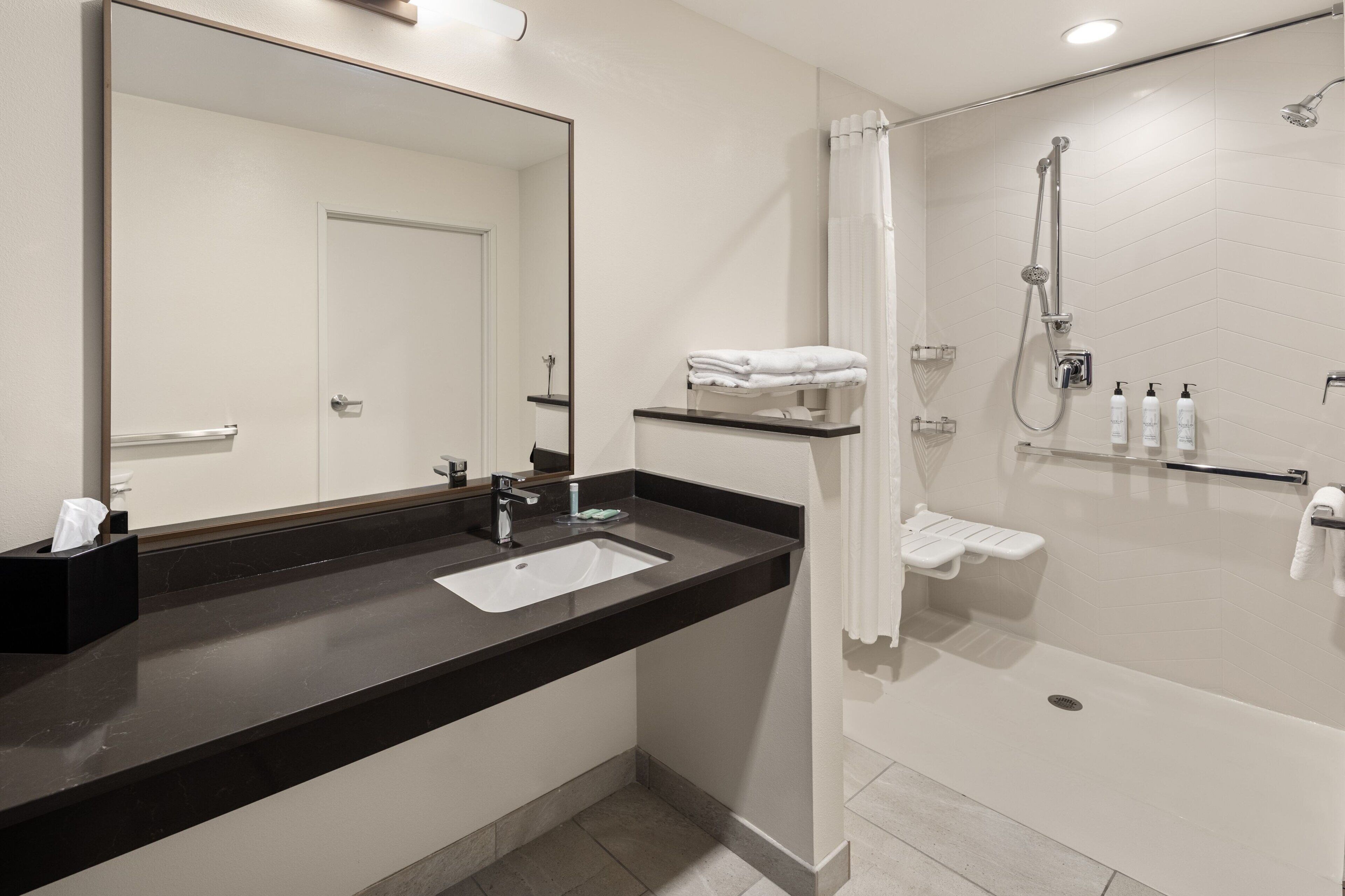 Un baño moderno con lavamanos de encimera negro, un espejo grande, ducha con cabezal de ducha manual y un perchero.
