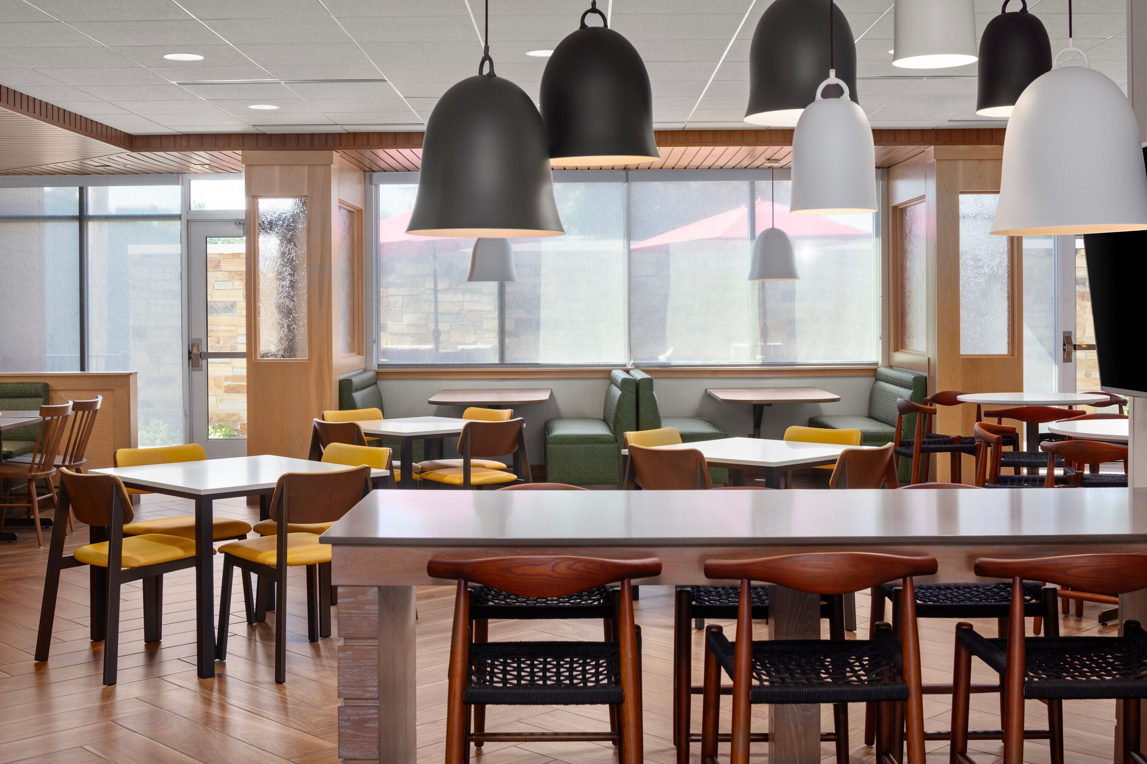 Un restaurant avec des tables et des chaises en bois, des suspensions noires et blanches et de grandes fenêtres.