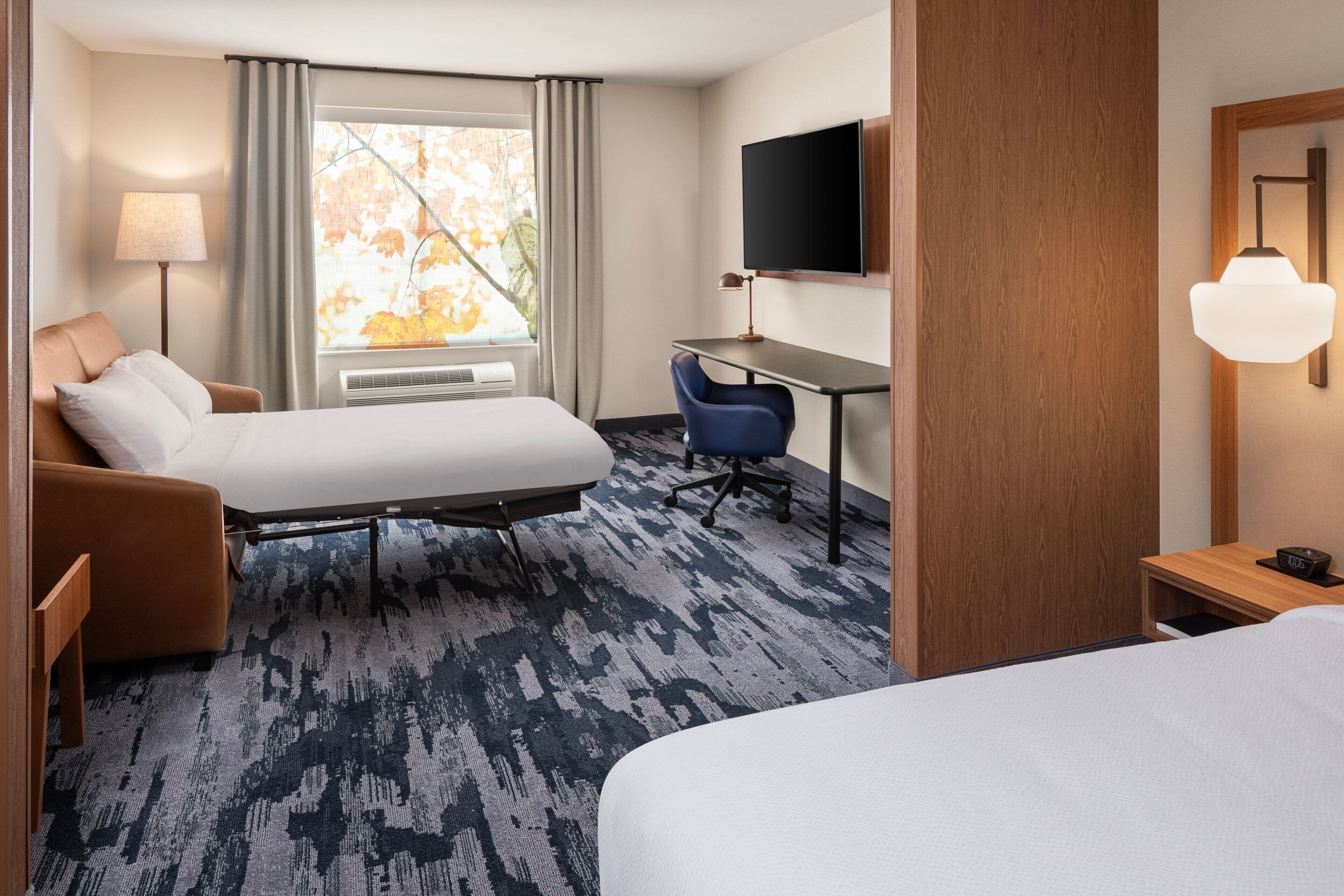 A hotel room with a bed, a desk, a chair, a TV, and a window with curtains.