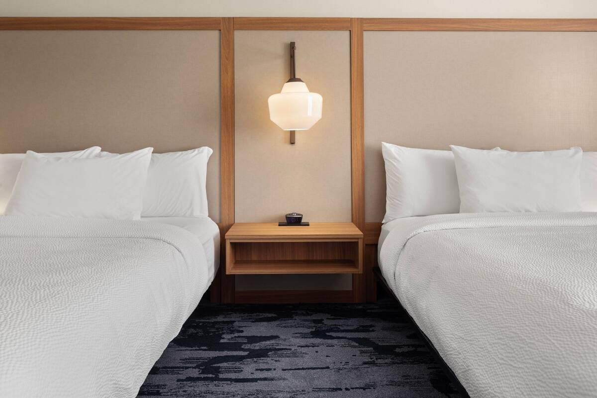 A hotel room with two beds, a nightstand with a clock, and a wall-mounted light fixture.