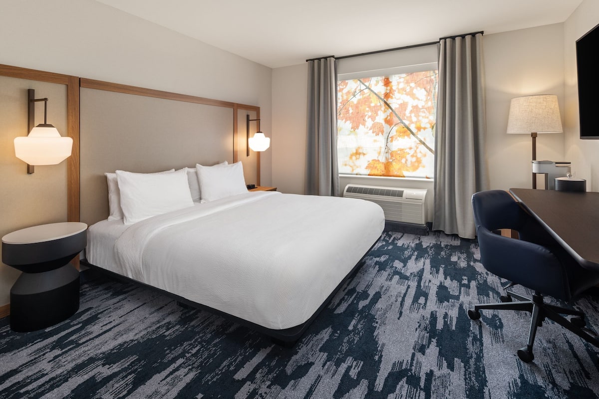 A hotel room with a large bed, a desk, a chair, a round side table, a wall-mounted lamp, a window with curtains, and a wall-mounted air conditioning unit.
