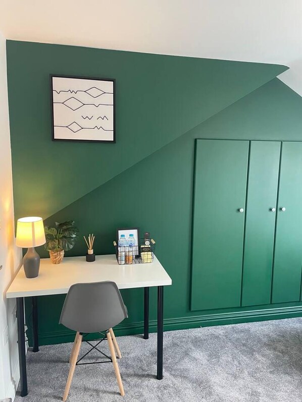 Une pièce avec un mur d’accent vert, un bureau avec une lampe, une chaise, un tableau encadré et un meuble.