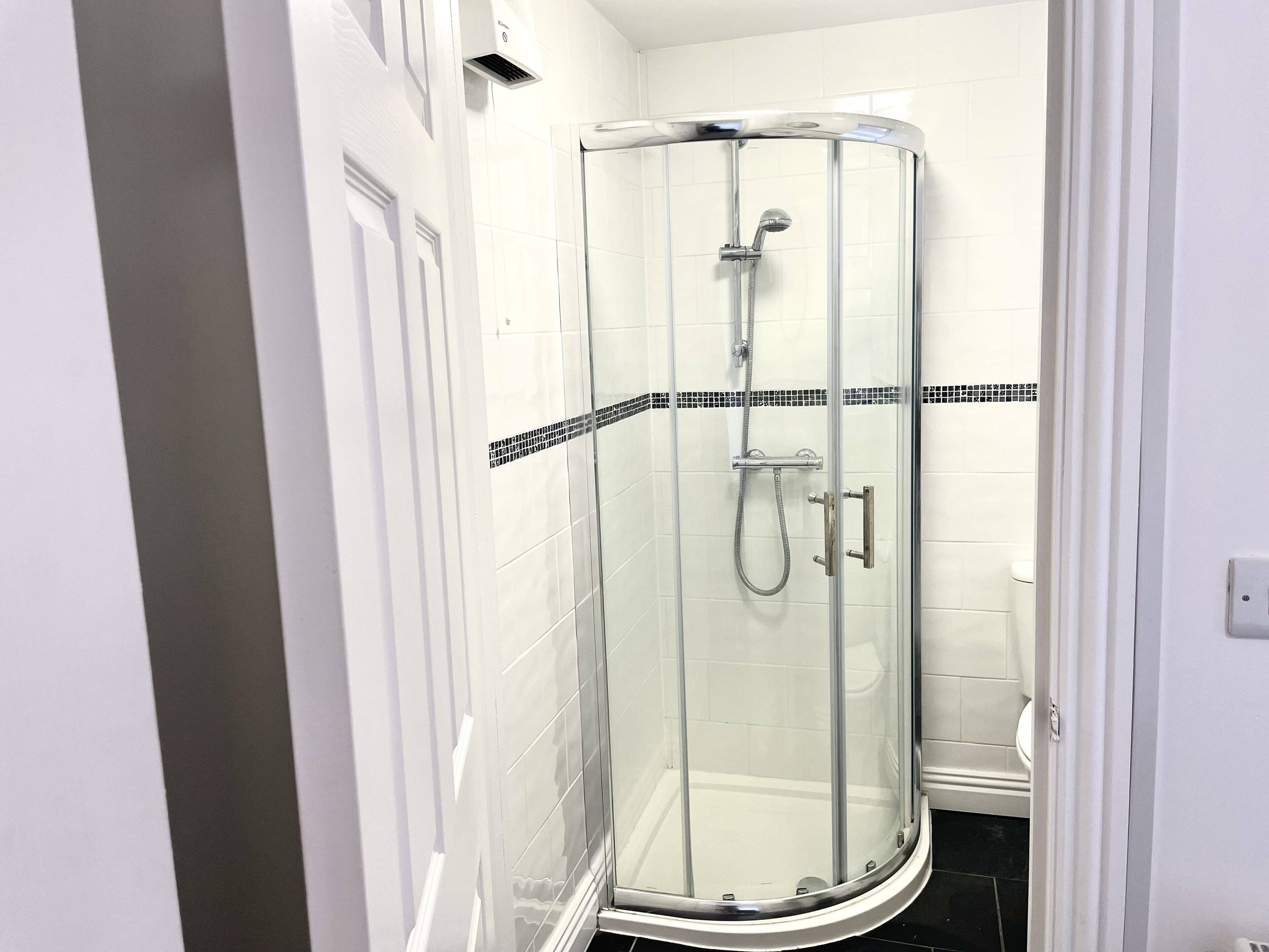 Une douche fermée par des parois de verre, dotée d’un pommeau de douche et d’un flexible à main, de murs recouverts de carreaux blancs et d’une bordure de carreaux noirs et blancs.