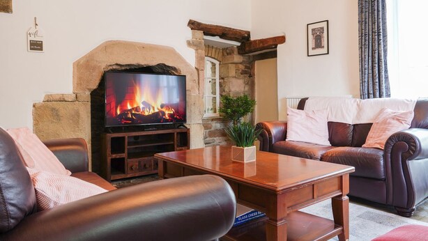 A cozy living room with a stone fireplace, leather sofas, a wooden coffee table, and a television displaying a fireplace scene. - The Old Bulls Head (High Peak)