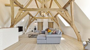 A spacious loft with exposed wooden beams, a grey sectional sofa, and a fireplace.