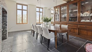 A dining area with a large wooden table, six chairs, a vase with flowers, and a brick fireplace.
