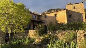Um edifício rústico de pedra com telhado de telhas, rodeado por vegetação e um muro de pedra.
