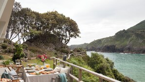 Ein Essbereich im Freien mit gedecktem Tisch, mit Blick auf eine Küstenlandschaft mit Hügeln und einem Gewässer.