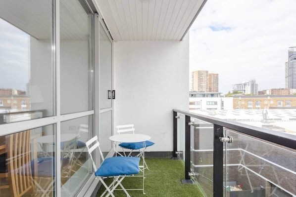 A balcony with a glass wall, a small table with chairs, and a view of the cityscape.