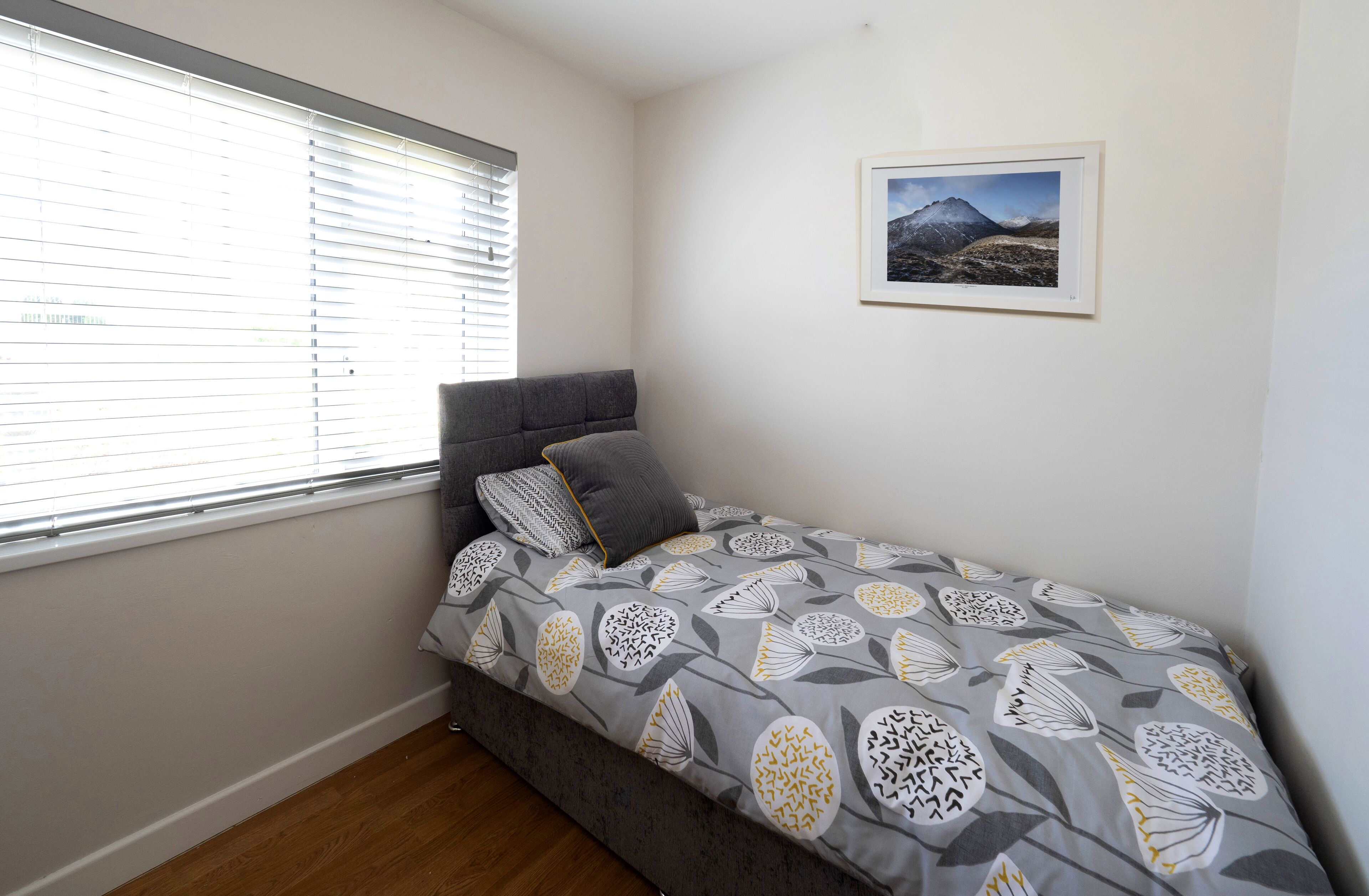 Une chambre avec un lit simple, une tête de lit grise, une fenêtre avec des stores et un tableau encadré au mur.