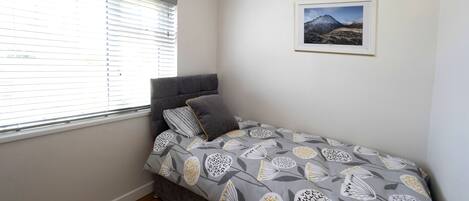 Une chambre avec un lit simple, une tête de lit grise, une fenêtre avec des stores et un tableau encadré au mur.