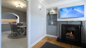 A modern living room with a fireplace, a television mounted above it, and a kitchen visible through an open door. - Charming 4-bed Cottage in Dungannon (Dungannon)