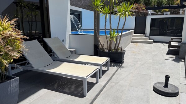 Uma área de estar moderna ao ar livre, com espreguiçadeiras, piscina e plantas em vasos.