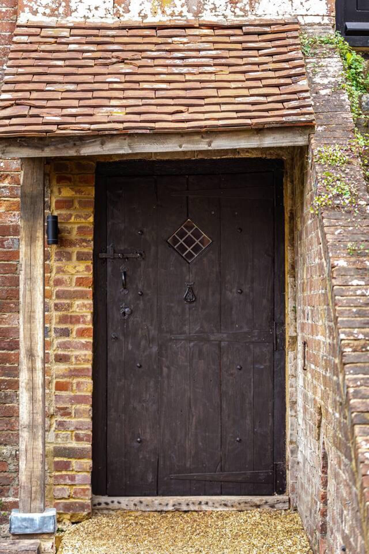 A dark wooden door with a diamond-shaped window and a small triangular emblem, set in a brick wall with a tiled roof above.