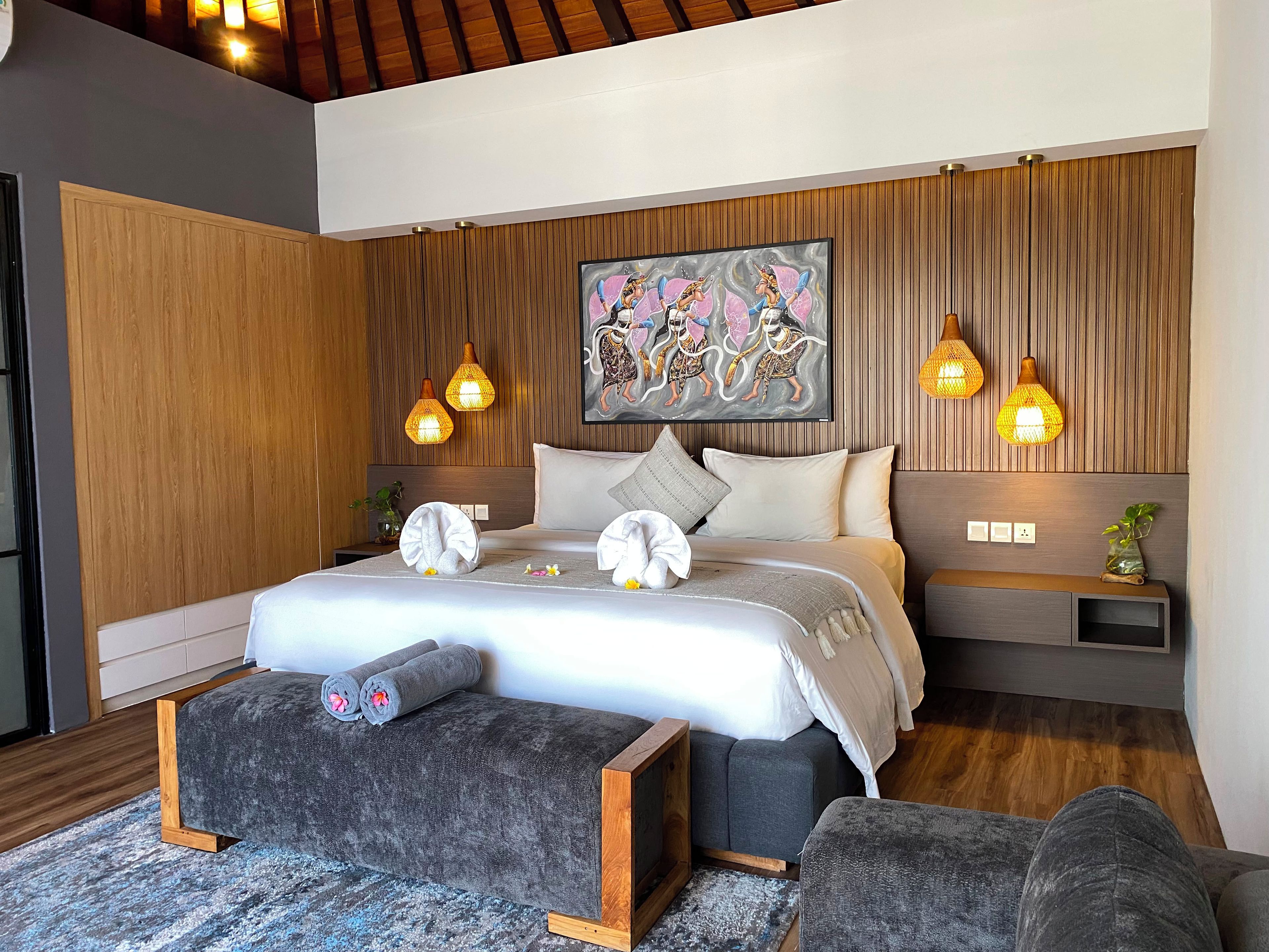 A modern bedroom with a large bed, wooden paneling, a painting, and pendant lights.
