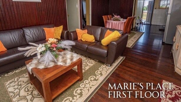 Una sala de estar con un sofá de cuero marrón, una mesa de centro de madera con un arreglo floral, y un comedor con mantel rosa.