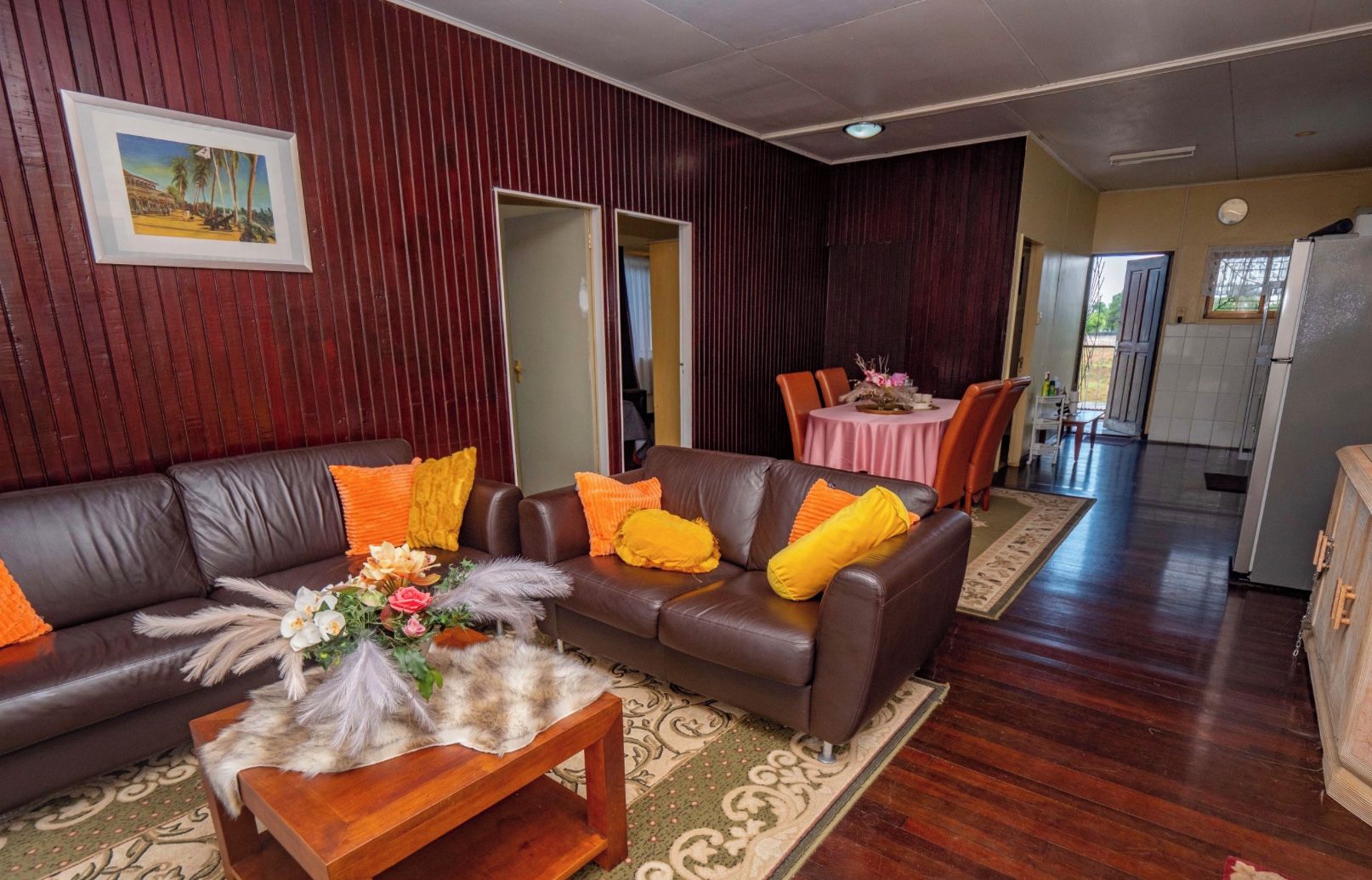 A living room with a brown leather sofa, a wooden coffee table with a floral arrangement, and a dining area with a pink tablecloth.