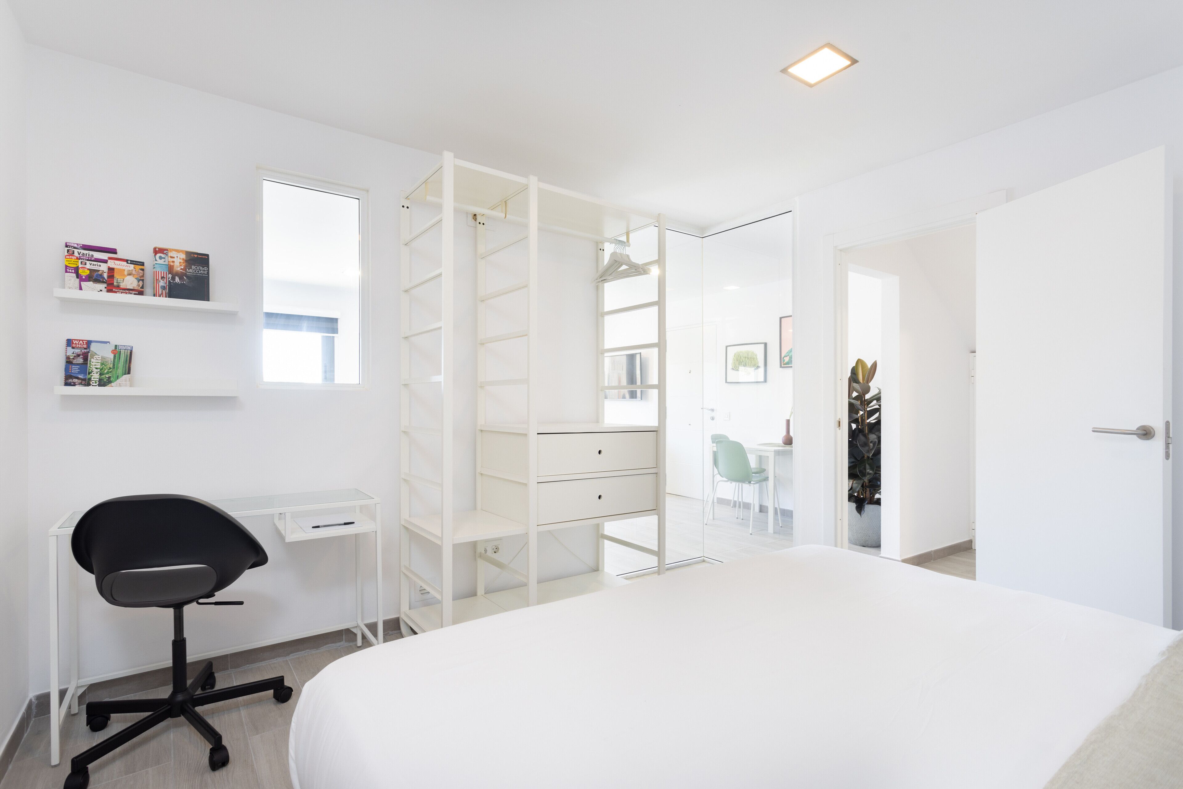A bedroom with a bed, a desk, and a chair. There is a wardrobe with shelves and a door.