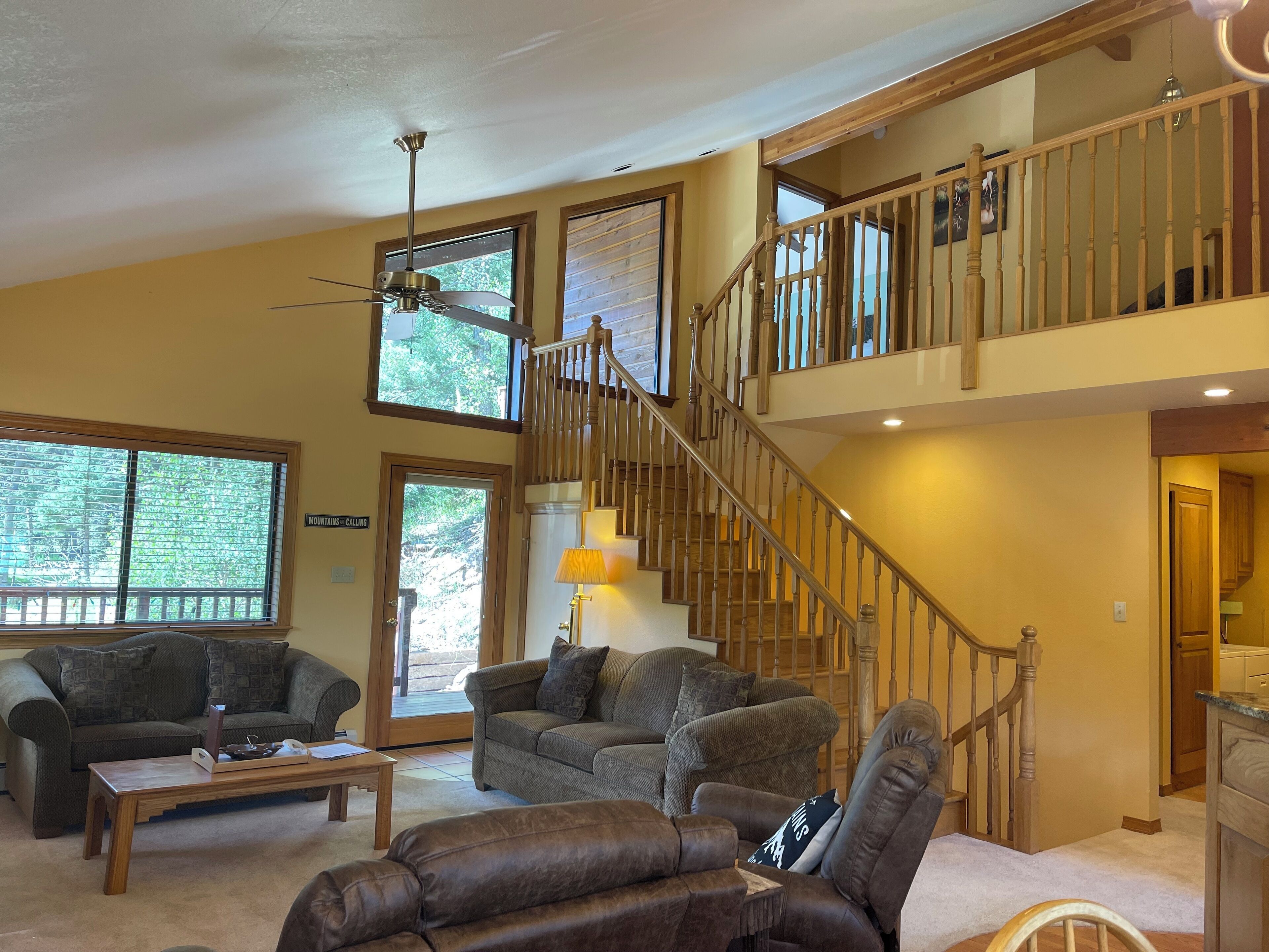 A spacious living room with a high ceiling, a wooden staircase, and large windows offering a view of the outdoors.