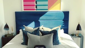A modern bedroom with a large abstract painting, a blue headboard, and multiple pillows. - Stylish and Chic 1 Bedroom Apartment in Canning Town (London)
