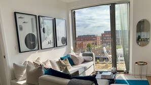 A modern living room with a sofa, a coffee table, and a view of the city through large windows. - Stylish and Chic 1 Bedroom Apartment in Canning Town (London)