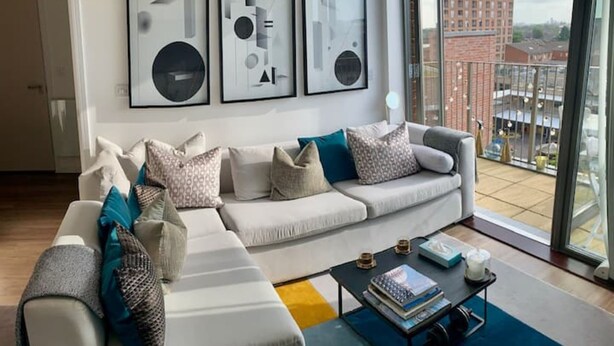 A modern living room with a white sectional sofa, a coffee table, and a view of the city through large windows. - Stylish and Chic 1 Bedroom Apartment in Canning Town (London)