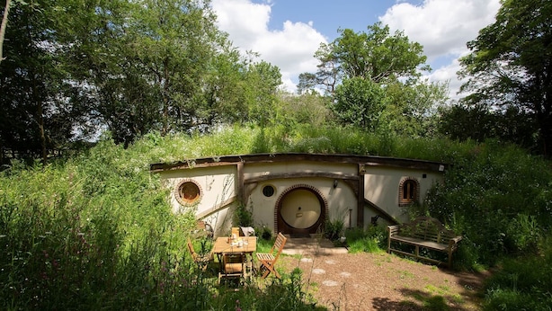 Un edificio rústico de piedra con puerta y ventanas redondas, rodeado de abundante vegetación.