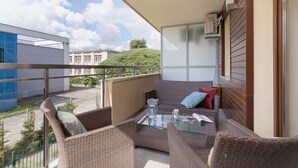 A balcony with wicker furniture, a glass table, and a view of buildings and greenery.