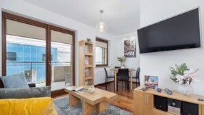 A modern living room with a sofa, coffee table, and TV. There's a dining area with a table and chairs, and a bookshelf.