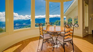 A dining area with a large round table and six chairs, large windows with a view of mountains and clouds, and wooden flooring.