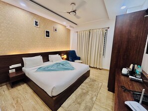 A hotel room with a large bed, a bedside table, a chair, a desk, a ceiling fan, and a window with curtains. - Bangalore Times by Alaya Stays (Bengaluru)