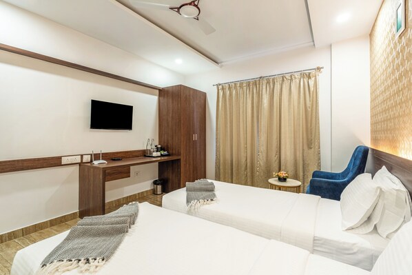 A hotel room with two beds, a desk with a flat-screen TV, a ceiling fan, and a window with curtains. - Bangalore Times by Alaya Stays (Bengaluru)