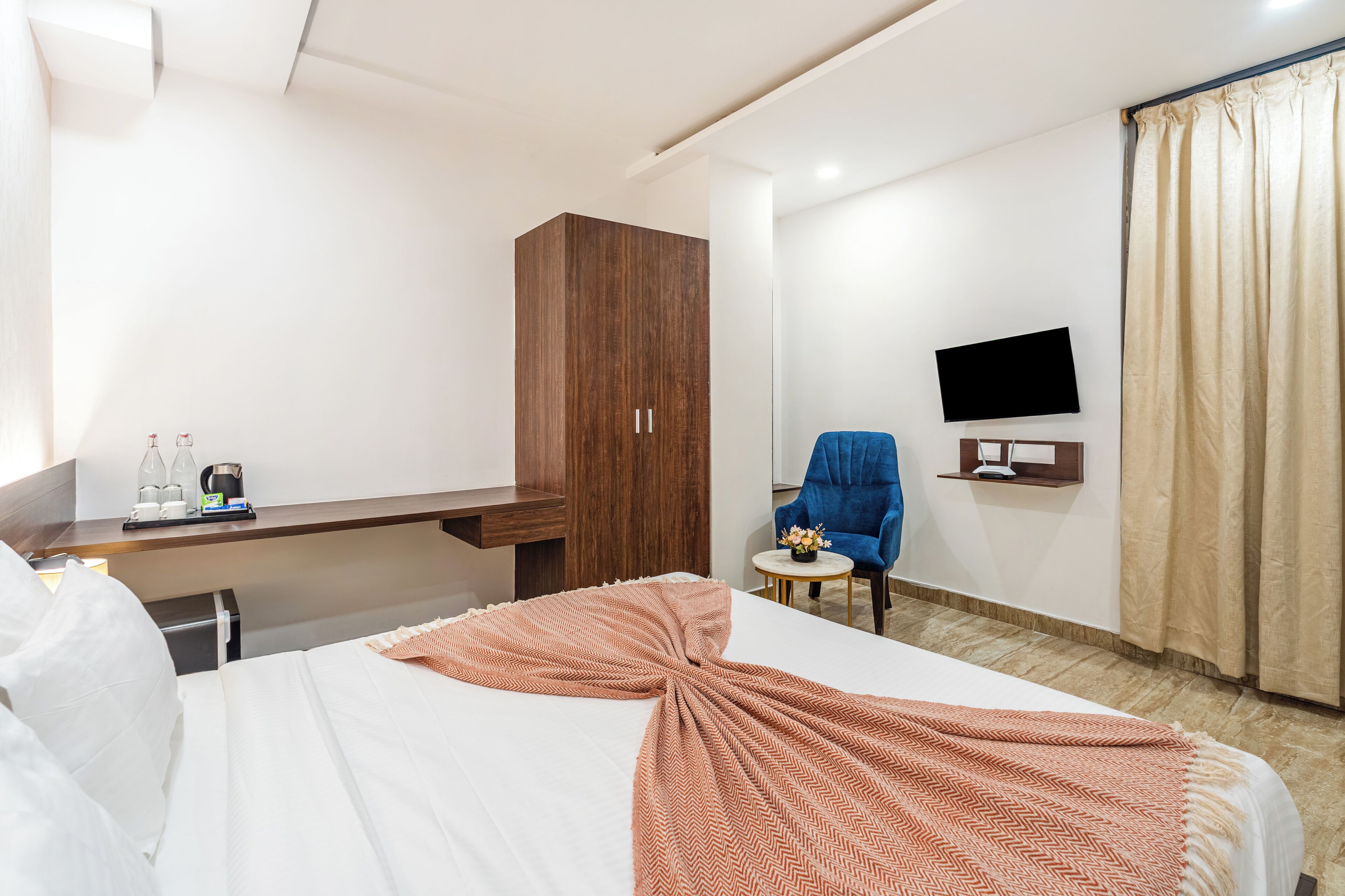 A hotel room with a bed, a wooden wardrobe, a desk with a coffee maker, a chair, a TV on the wall, and a curtain.