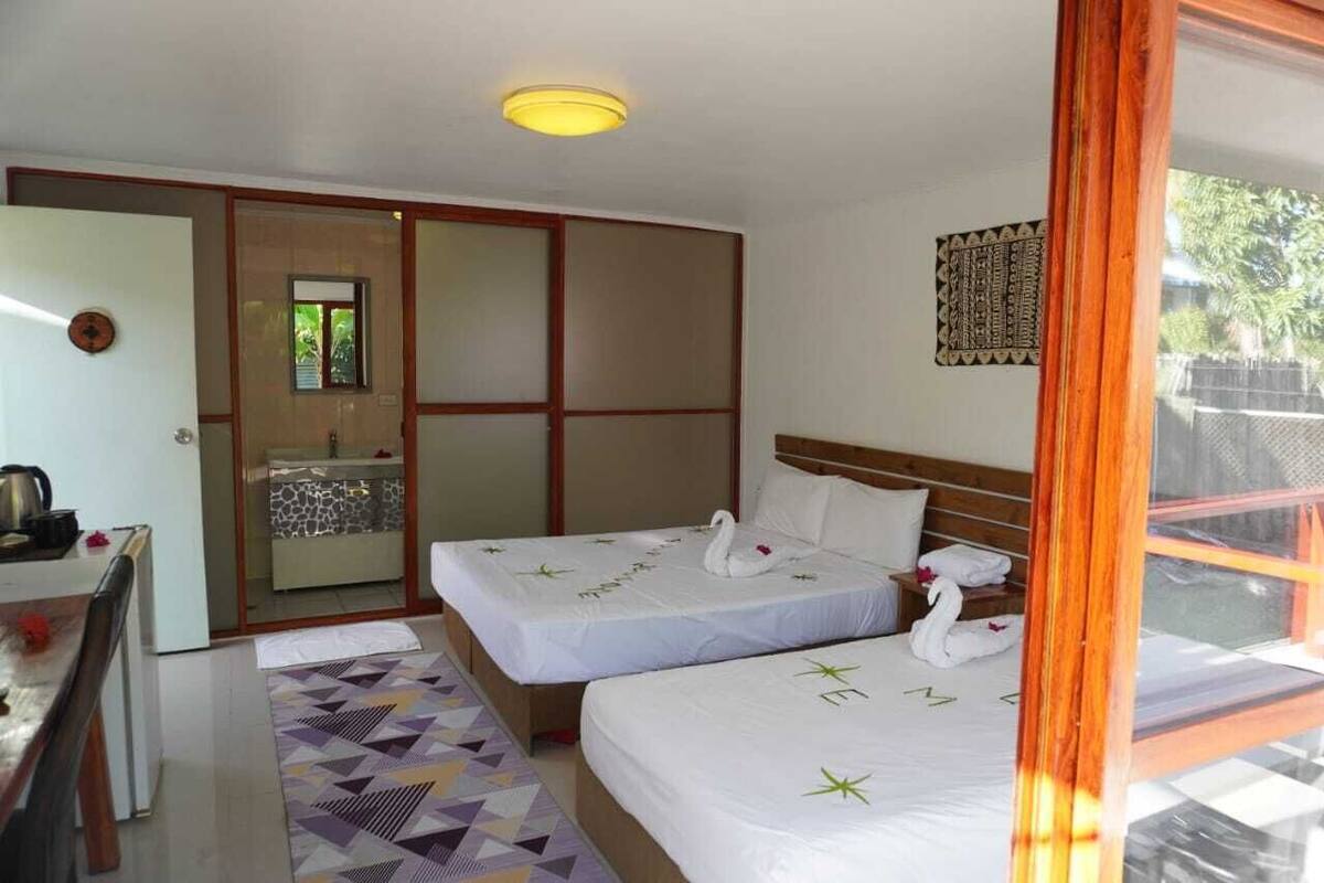 A hotel room with two beds, a small kitchenette, a bathroom with a glass door, and a view of greenery outside.