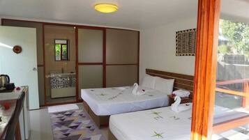 Quarto de hotel com duas camas, uma kitchenette pequena, banheiro com porta de vidro e vista para o verde lá fora.