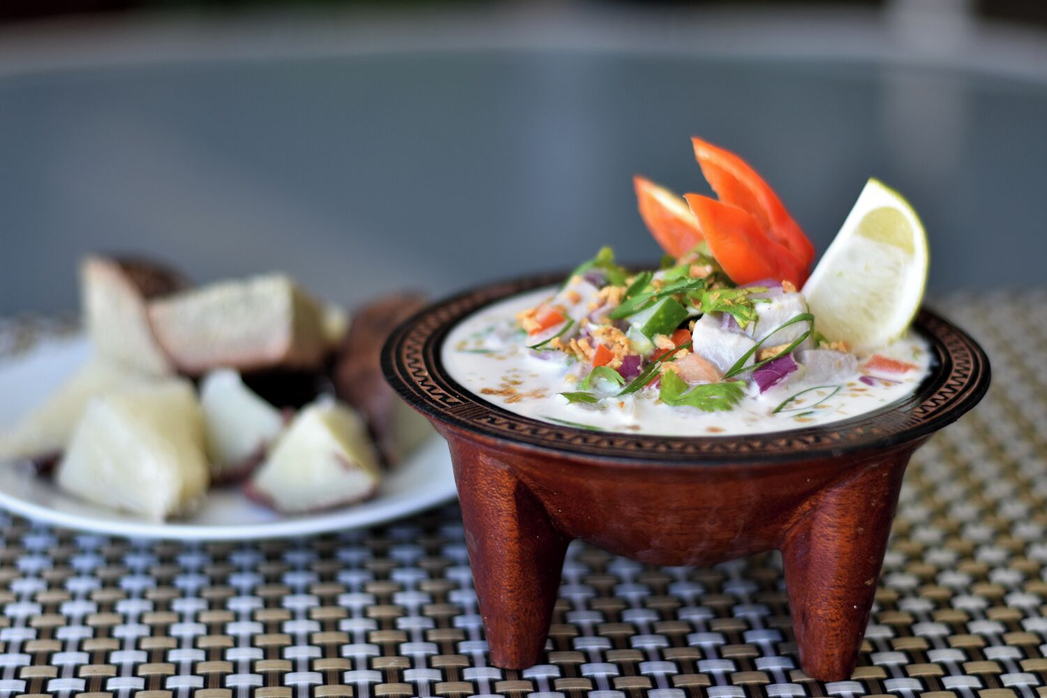 A bowl of soup with vegetables and a lemon wedge on a woven tablecloth.