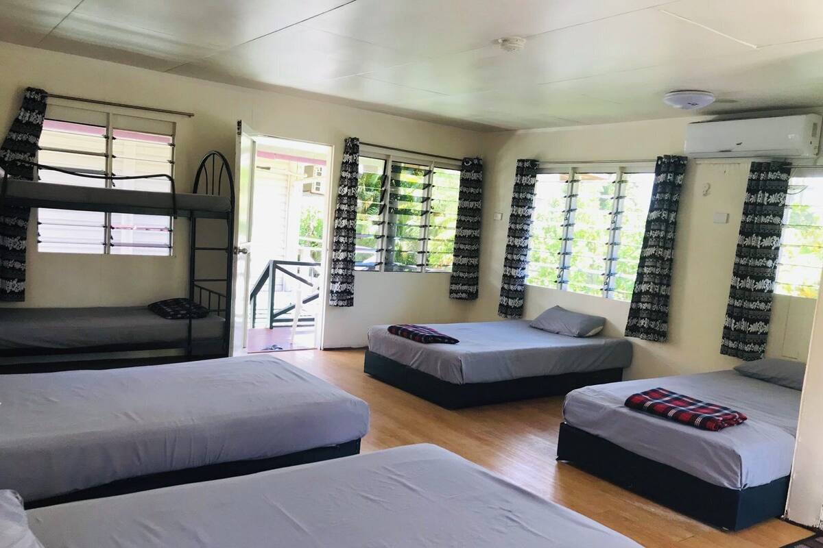 A dormitory room with bunk beds, two single beds, and a staircase leading to an outdoor area.