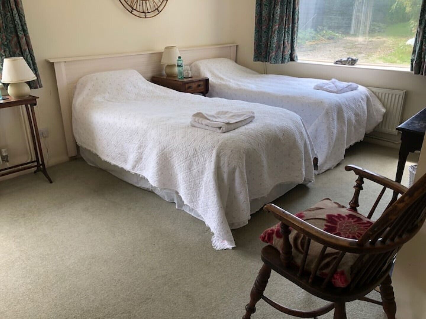 Une chambre avec deux lits simples, une table de chevet avec une lampe, une chaise et une vue sur l’extérieur par une fenêtre.