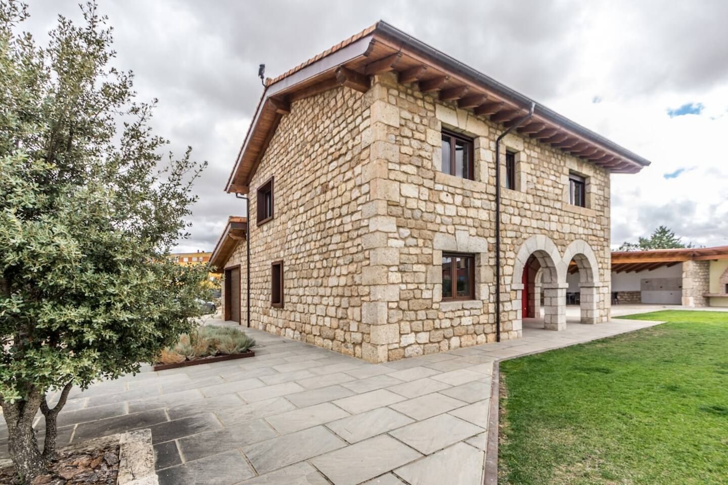 Un edificio de piedra con techo plano, entrada empavedada y un área verde al frente.