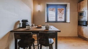 A small kitchen with a wooden table, two chairs, a coffee maker, cups, and a plate of croissants. There is a window with a view of a building outside. - Fidalsa Rest Station (Villadiego)