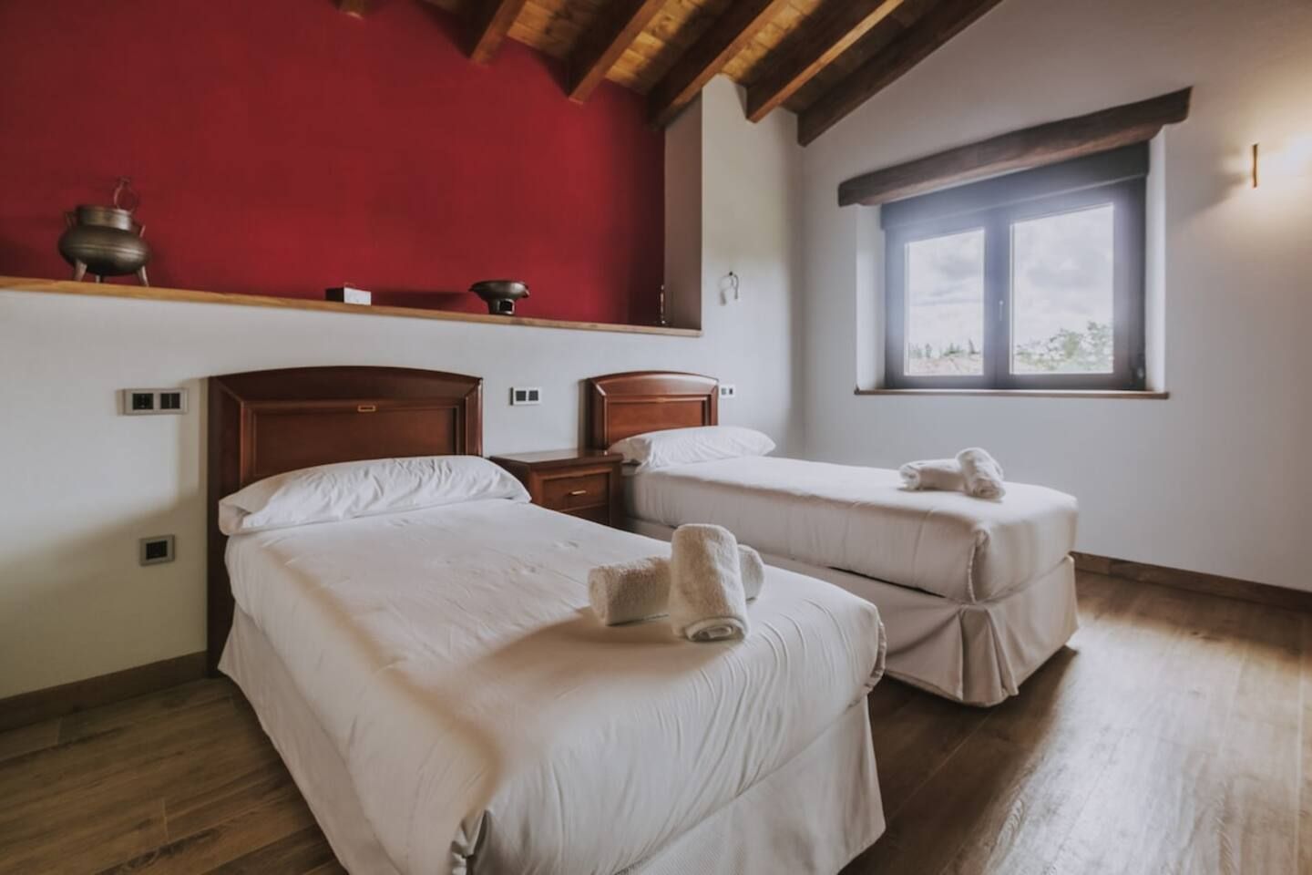 Una habitación con dos camas, una pared roja, una ventana y techo de madera.