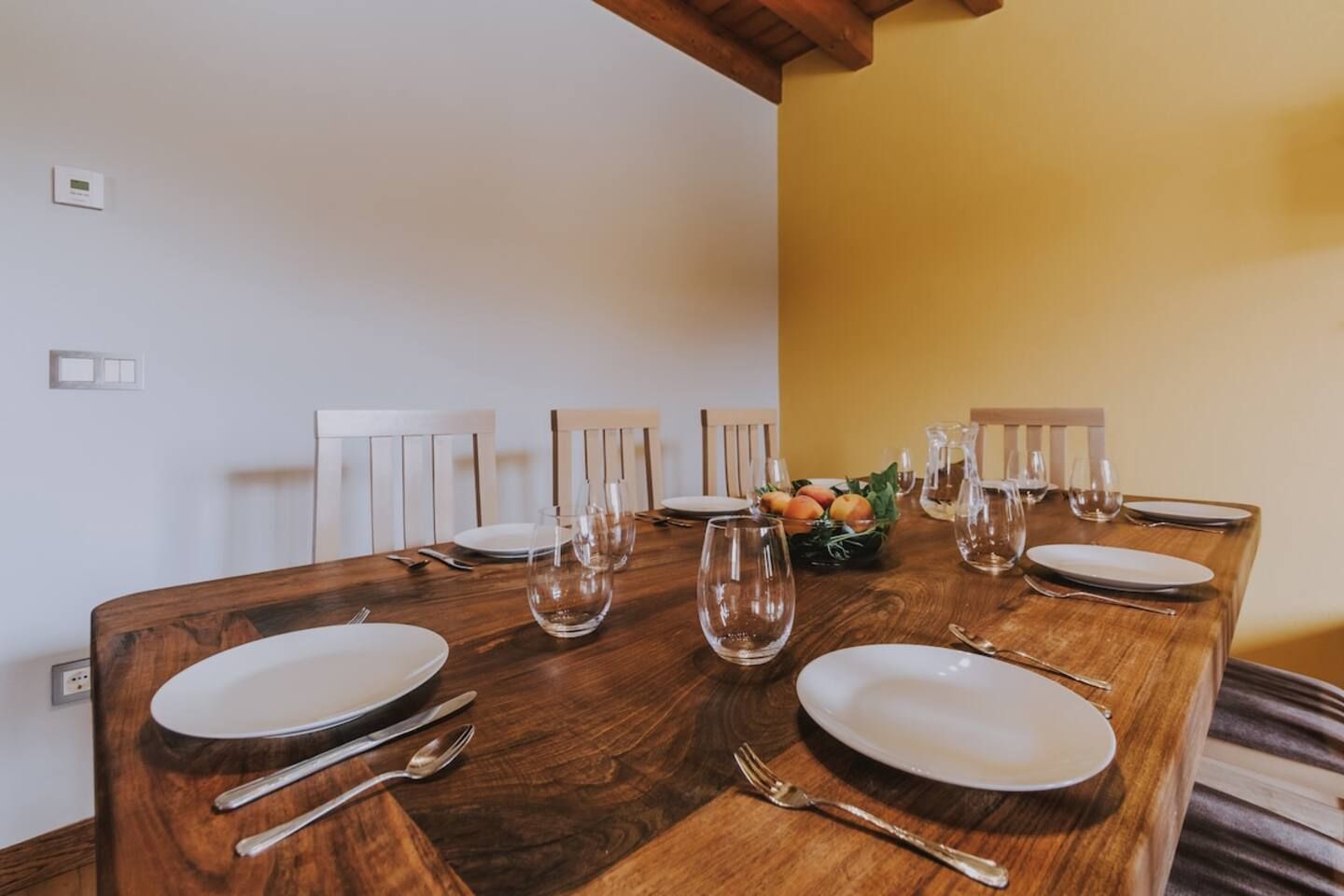 Una mesa de comedor dispuesta con platos, cubiertos y vasos, con un frutero sobre la mesa.