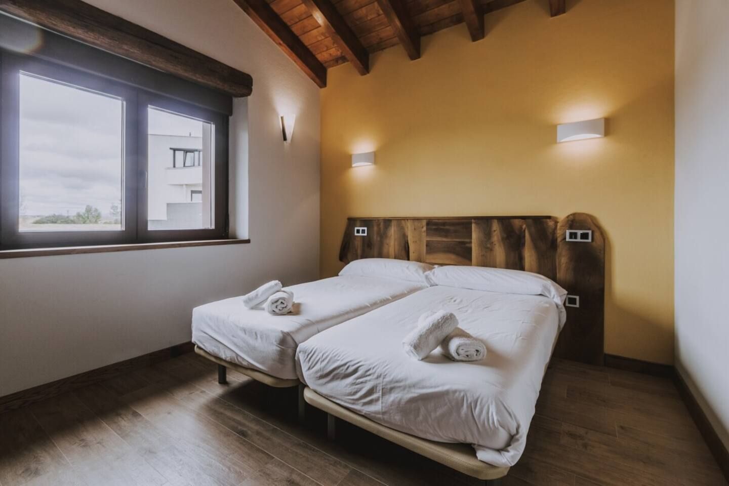 A room with two beds, wooden headboards, a window, and a view of a building outside.
