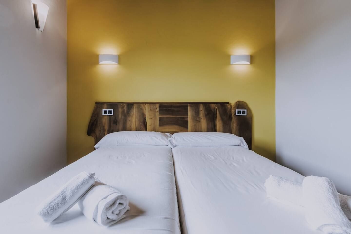 Cama doble con ropa blanca y dos lámparas fijadas a la pared.