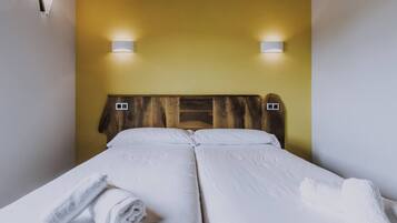 Cama doble con ropa blanca y dos lámparas fijadas a la pared.