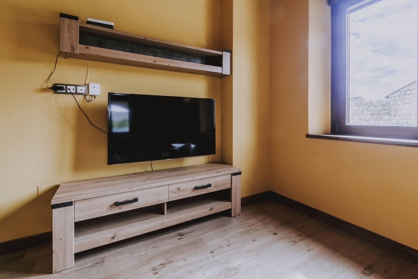 Habitación con televisión en mueble de madera, ventana con vista a pared de piedra y toma de corriente en la pared.